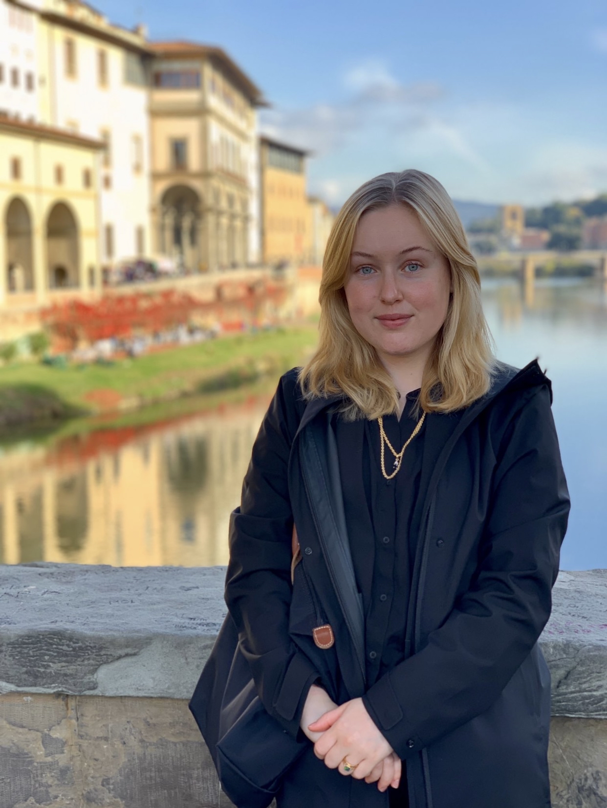 Former Human Rights Law Clinic student and LLM graduate Emma Lennhammer leaning against a low wall, with a background of blue sky reflected in the water of a river running by buildings with arched architecture and flowers.